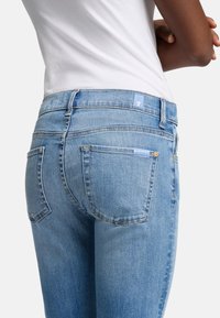 Jean en denim bleu clair avec une coupe ajustée, comportant deux poches arrière et une petite étiquette bleue sur la taille. Texture lisse.