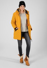 Gelbe Kapuzen-Puffjacke mit gestepptem Muster, grauer Pullover, dunkle Jeans und braune Stiefeletten mit Fellfutter und Riemen.
