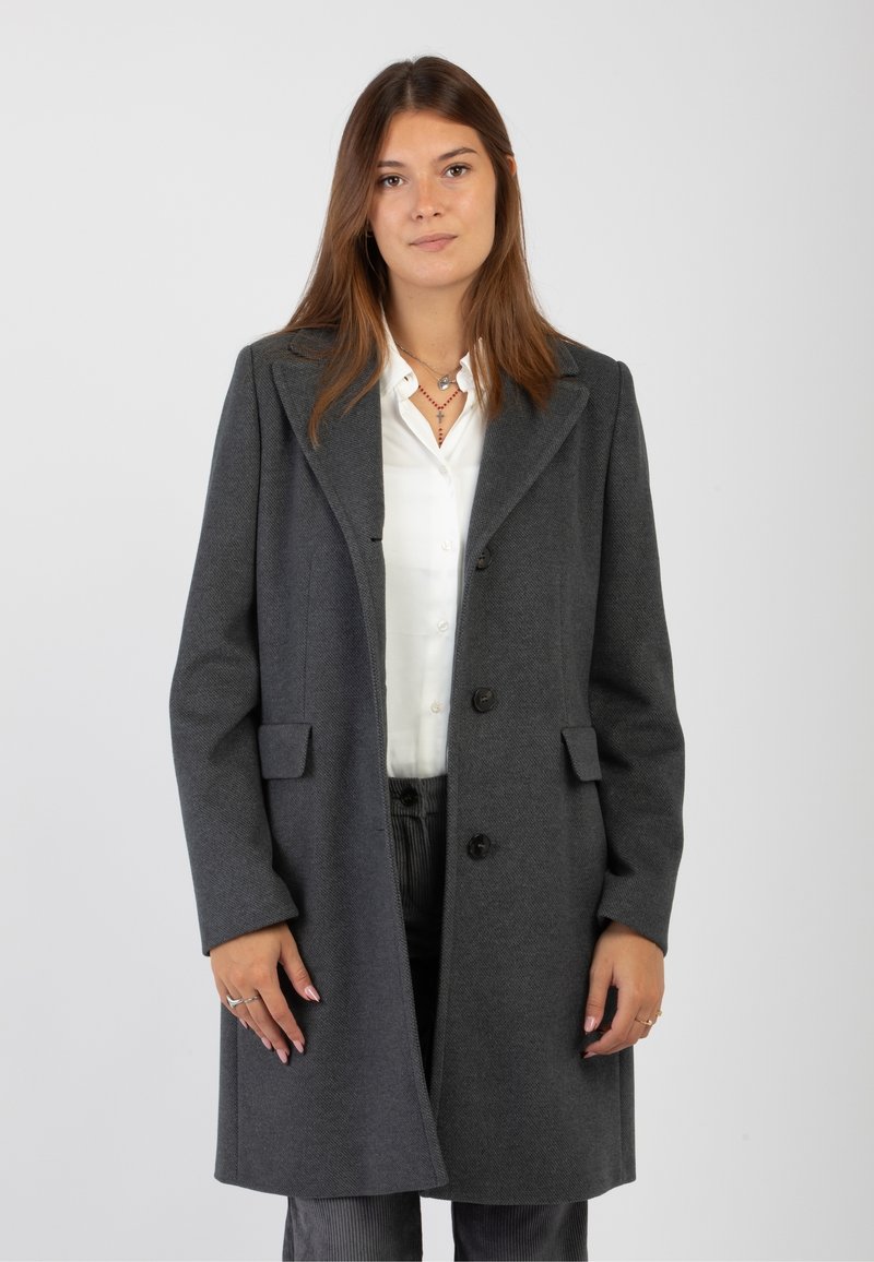 Giovane donna che indossa un cappotto grigio scuro sopra una camicia bianca, in piedi contro uno sfondo chiaro e uniforme, che guarda la telecamera con un'espressione neutra.
