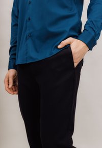 Teal skjorte med knapper laget av glatt stoff, brukt med mørke, ledige bukser. Skjorten har avrundet kant, og en hånd er i bukselommen.