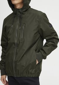 Olive green hooded jacket with a zip front, elastic cuffs, side pockets, and a small logo patch on the sleeve. Lightweight, smooth texture.