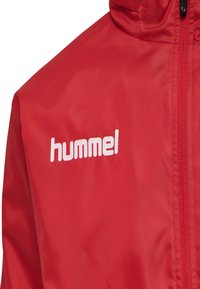 Giacca rossa realizzata in materiale liscio e leggero, con zip completa e logo bianco sulla parte sinistra del petto. Dettagli di cucitura in evidenza.