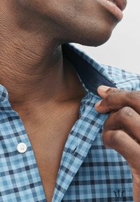 Nærbillede af en person, der justerer kraven på en blå- og sortternet skjorte, hvilket fremhæver hudens struktur og håndbevægelse.