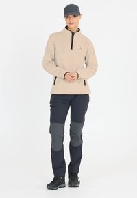 Beige fleece pullover met halve rits en zwarte accenten, gecombineerd met donkere broek met grijze kniebeschermers en ritszakken. Zwarte schoenen en pet.