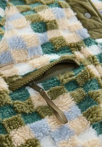 Jachetă din fleece cu un model multicolor în carouri, în nuanțe de verde, bej și albastru; include o bivă cu fermoar și un tab de țesătură.