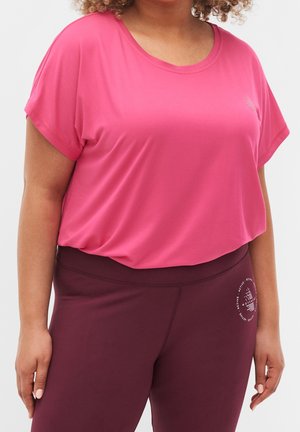 Woman wearing a loose pink short-sleeve top and high-waisted maroon leggings with a circular logo on the left hip.