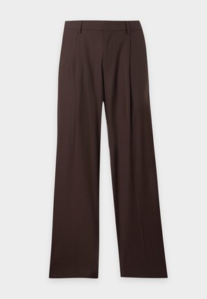 Homme portant un t-shirt violet, un pantalon large marron foncé, des baskets blanches et des lunettes de soleil teintées en bleu, debout sur fond blanc uni.