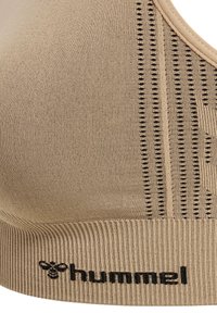 Close-up van een beige geribde sportbeha met zwarte verticale sierlijke strepen en het "hummel" logo op de band.