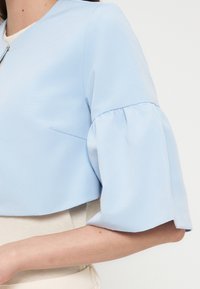 Femme portant un chemisier bleu clair avec des manches bouffantes jusqu'au coude et un col rond, associé à un pantalon de couleur claire.