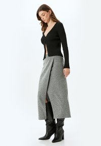 Cardigan noir à côtes avec un décolleté en V profond, associé à une jupe portefeuille grise avec une fente sur le côté. Le mannequin porte des bottes noires à talons montant au genou.