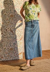 Grøn blomstret kortærmet bluse parret med en lang lyseblå denimskørt og beige slip-on sko, sat mod en mønstret baggrund.