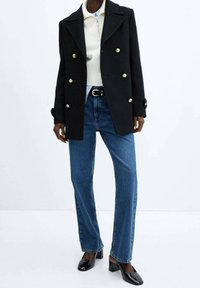 Abrigo negro cruzado con botones dorados, suéter blanco con textura acanalada, jeans azules de pierna recta y zapatos de tacón cuadrado de charol negro.