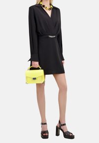 Svart omlottklänning med långa ärmar, med en kedjedetalj i midjan. En gul handväska och svarta platåsandaler fulländar looken.