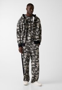 Completo felpa e pantaloni con motivo mimetico in tonalità di grigio e nero. Felpa con cappuccio, zip frontale, polsini a coste e logo sul petto.