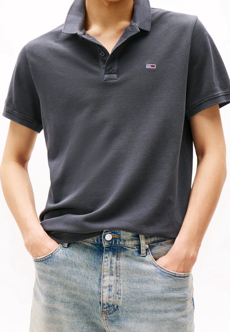Polo gris foncé en tissu texturé avec des manches courtes, arborant un petit logo. Associé à un jean en denim bleu clair.