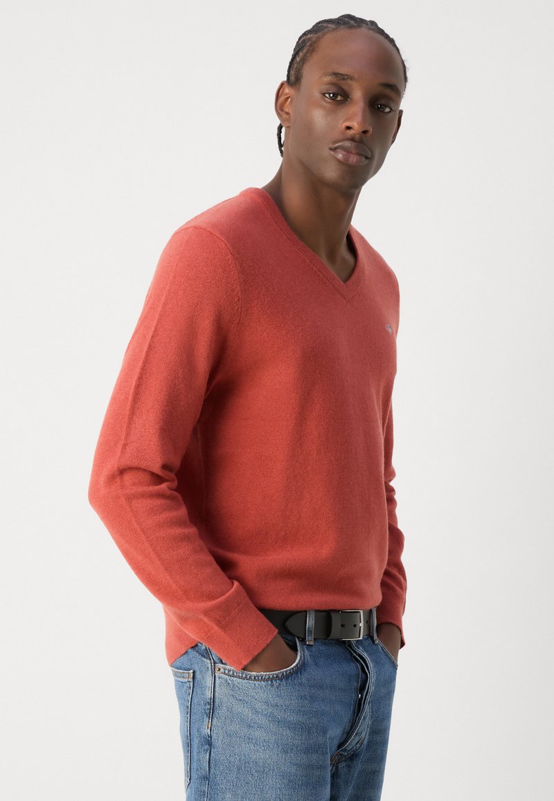 V-Ausschnitt-Pullover in Ziegelrot, aus weichem Stoff gefertigt. Slim Fit mit langen Ärmeln, kombiniert mit blauen Jeans und einem schwarzen Gürtel.
