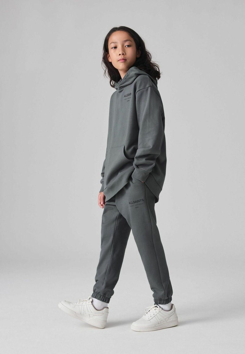 Siv hooded sweatshirt in mehki tkanini in ujemajoči se jogger hlače, ki vsebujejo logotip blagovne znamke v črni barvi. Bele superge dopolnjujejo outfit.
