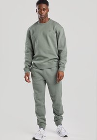 Felpa in pile verde oliva e pantaloni coordinating con polsini a coste. Presenta un logo minimalista sul petto ed è abbinata a sneaker bianche.