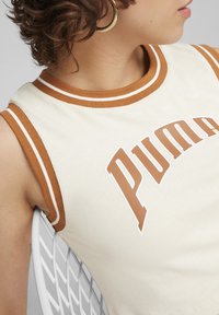 Top senza maniche crema con scollatura e bordi delle maniche in maglia marrone, caratterizzato da un grande logo bianco "PUMA" in carattere marrone sul petto.
