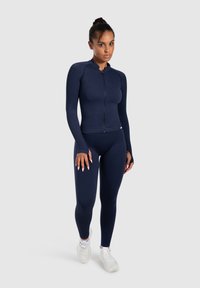 Top de manga larga con cremallera en color navy, combinado con leggings ajustados a juego. Tela suave y elástica con una textura lisa. Calzado deportivo ligero.