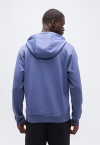 Homme portant un sweat à capuche bleu, vu de dos sur un fond clair uni.