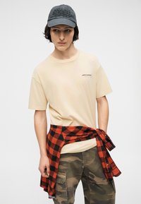 Νεαρός που φοράει μπεζ μπλούζα McLaren, μαύρο και κόκκινο καρό πουκάμισο δεμένο γύρω από τη μέση, παραλλαγής cargo παντελόνι και σκούρο γκρι καπέλο.