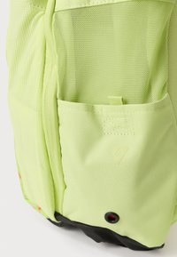 Mochila de tela verde claro con bolsillo de malla, pequeño bucle cosido y parte inferior negra con un orificio de ventilación redondo acentuado en rojo.