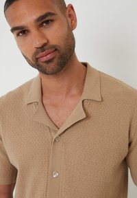 Camisa beige de manga corta con un patrón texturizado, cuello con muesca y dos botones en el frente. La tela parece suave y ligera.