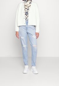 Vit rippad cardigan med rund halsringning och knappar, parat med ljusblå rivna jeans och vita sneakers, som står framför en neutral bakgrund.