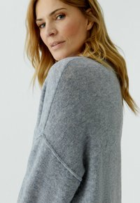 Vrouw met lang blond haar en een grijze gebreide trui, kijkt over haar schouder tegen een effen lichte achtergrond.