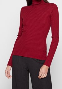 Pull à col roulé côtelé rouge à manches longues, avec une coupe ajustée et une texture lisse. Associé à un pantalon noir texturé.