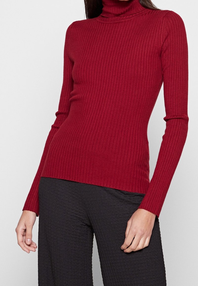 Pull à col roulé côtelé rouge à manches longues, avec une coupe ajustée et une texture lisse. Associé à un pantalon noir texturé.