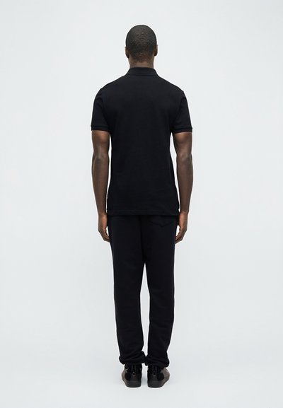 Άνδρας που στέκεται με την πλάτη γυρισμένη, φορώντας μαύρο πόλο με κοντά μανίκια, μαύρο παντελόνι και μαύρα αθλητικά παπούτσια μπροστά σε απλό ανοιχτό φόντο.