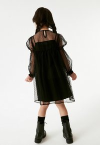 Schwarzes Polka-Dot-Kleid mit transparenten langen Ärmeln, einem Schlüsselrückenausschnitt und einem gerafften Taillenbereich. Getragen mit schwarzen Stiefeletten und passenden Socken.