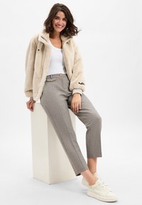 Lichtbeige, donzige jas met een rits, witte tanktop, grijze geruite broek met gouden knopen en witte sneakers, tegen een effen achtergrond.
