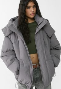 Graue Steppjacke mit hohem Kragen und großer Kapuze, mit Druckknöpfen und Steppdesign. Getragen über einem grünen Oberteil und Jeans.