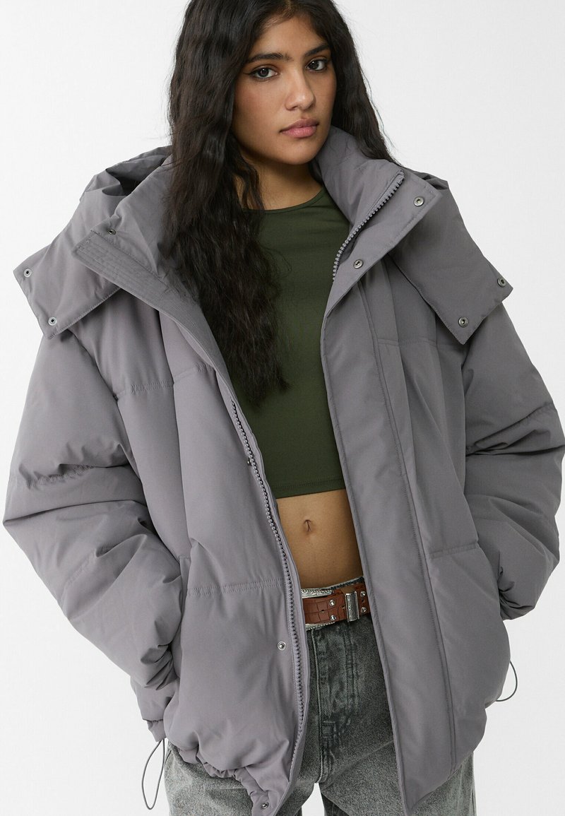 Graue Steppjacke mit hohem Kragen und großer Kapuze, mit Druckknöpfen und Steppdesign. Getragen über einem grünen Oberteil und Jeans.
