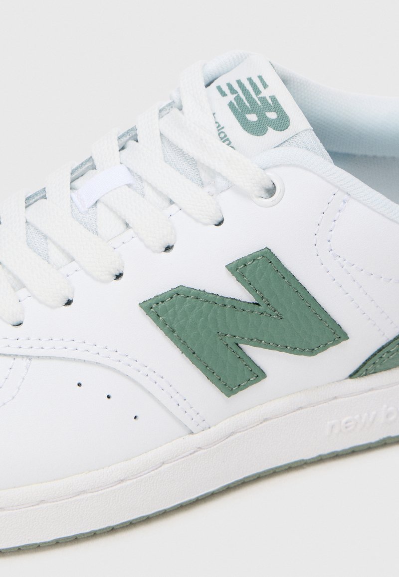 Zapatilla blanca de cuero con logo "N" texturizado en verde claro. Presenta cordones blancos y una lengüeta acolchada con detalle del logo. Suela de goma.