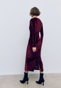 Burgundská sametová šaty s dlouhým rukávem, postranním rozparkem a řasením, doplněné černými kotníčkovými botami na podpatku.