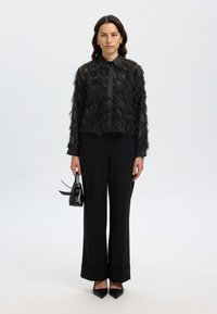 Black sheer blouse with feather-like texture and collar, paired with wide-leg black trousers and pointed-toe black heels. Holding a small black bag.
