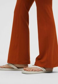 Roestkleurige flairbroek met contrasterende stiksels, gecombineerd met witte slippers. Focus op de stoftextuur en de bredere broekvorm.
