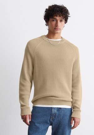 Beige Strickpullover mit Rippstruktur, Rundhalsausschnitt und langen Ärmeln, kombiniert mit blauen Jeans. Akzente setzen ein geschichtetes weißes Hemd.