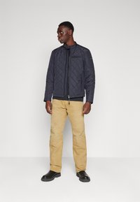 Jachetă din material quilted de culoare bleumarin, cu un model cusut în diagonală, închidere cu fermoar, buzunare laterale și guler căptușit, purtată cu pantaloni bej și cizme negre.