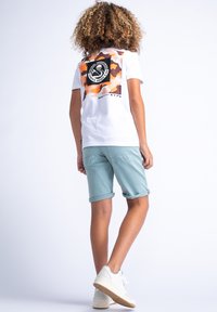 T-shirt bianca con design camouflage arancione sul retro, abbinata a pantaloni corti verde menta arrotolati in fondo e sneakers bianche con suole testurizzate.
