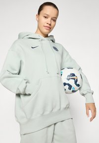Lys grønn hettegenser med lommer foran og snorer, med en liten navy Nike-logo. Personen holder en mønstret fotball.