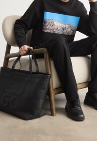 Černá kožená taška typu tote s raženým logem "HUGO", nošená s černým svetrem s barevným motivem a kombinovaná s černými kalhotami a botami.