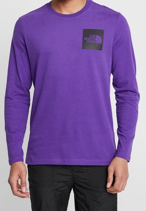 Mężczyzna w fioletowej koszuli z długim rękawem z czarnym kwadratowym logo North Face na lewej piersi i czarnych spodniach.