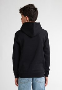 Felpa nera con cappuccio regolabile, texture morbida, polsini e orlo a costine. Indossata con jeans blu chiaro, che evidenziano un design casual.