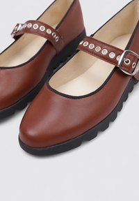 Zapatos Mary Jane de cuero marrón con punta redondeada, suela de goma negra y correa en el tobillo con ojales plateados y un hebilla de metal.