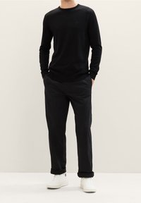 Maglione nero lavorato a maglia con scollo tondo, indossato con pantaloni neri e sneakers bianche. La vestibilità è ampia con polsini e orlo a coste.